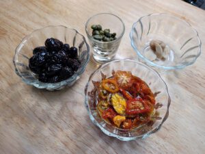 Dried tomatoes, olives, garlic, and capers in separate cups